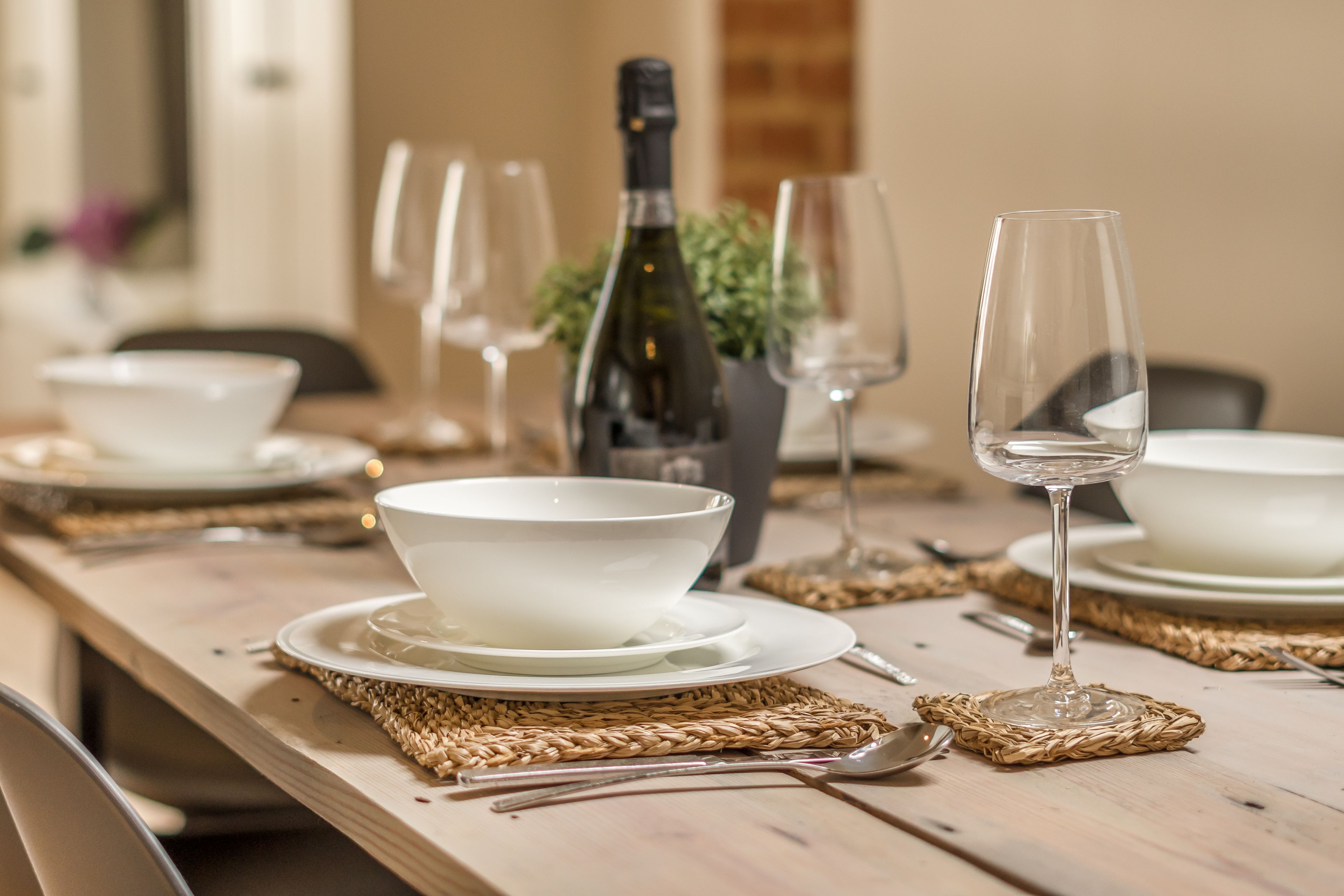 A dining table set with white bowls, plates, and wine glasses, with a bottle of wine and a potted plant in the background.