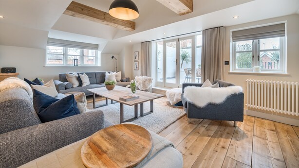 A spacious living room with a high ceiling, wooden beams, a large window, and a cozy seating area with a sofa and armchairs. - Burnham Loft - sleeps 4 guests  in 2 bedrooms (Burnham Market)