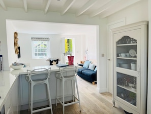 A modern kitchen with white bar stools, a dining table, and a sofa in the living area.