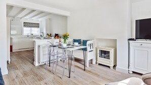 Una cocina moderna con armarios blancos, un comedor con mesa y sillas blancas, y un televisor pequeño sobre un mueble.
