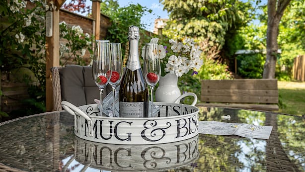 Uma mesa de vidro com uma chávena branca e uma garrafa de Prosecco, rodeada por uma cesta de vime com um menu.