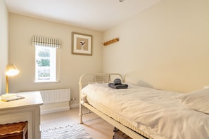 A bedroom with a bed, a desk with a lamp, a radiator, a window with curtains, and a framed picture on the wall. - Cherryhill - sleeps 5 guests  in 3 bedrooms (North Creake)