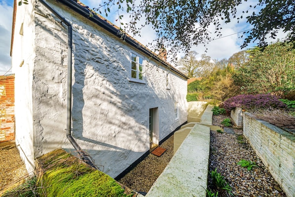 A white stone house with a small window, a door, and a pathway leading to a garden with gravel and plants.