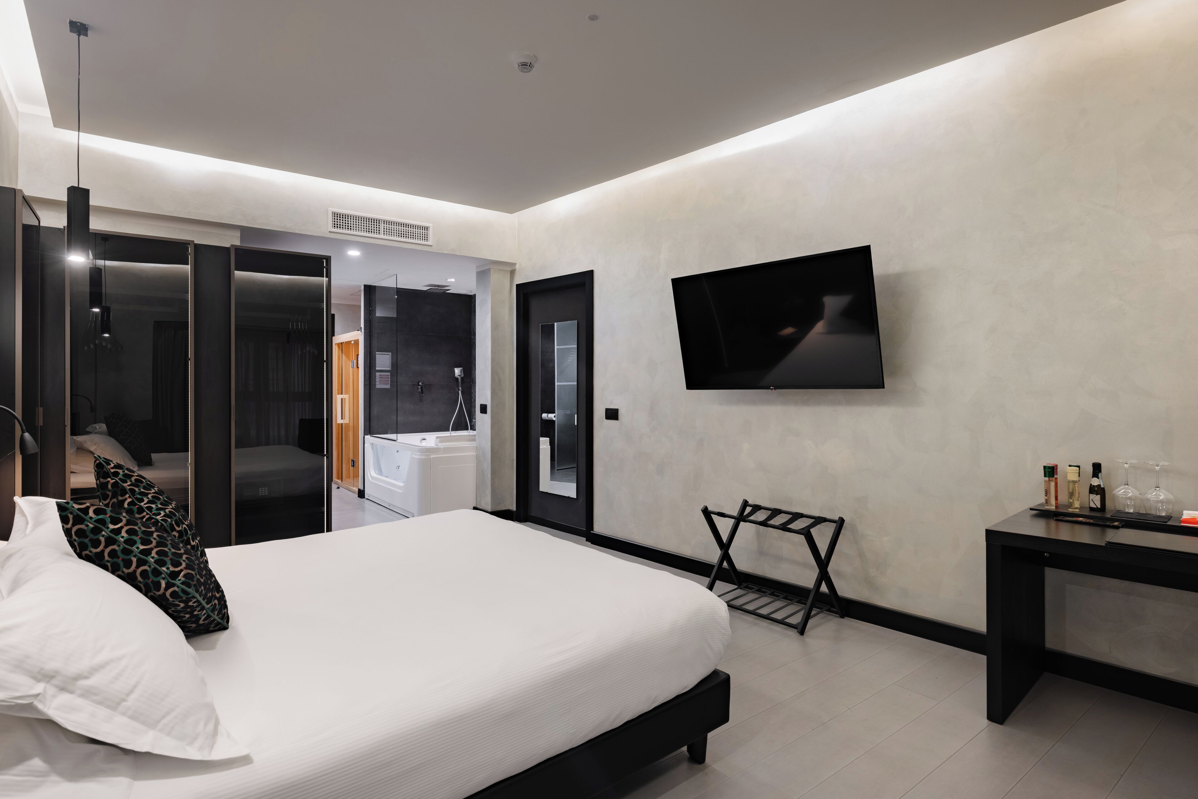 Una habitación de hotel moderna con una cama grande, un televisor de pantalla plana en la pared, una mesita con botellas y vasos, y un vestidor con puertas correderas.