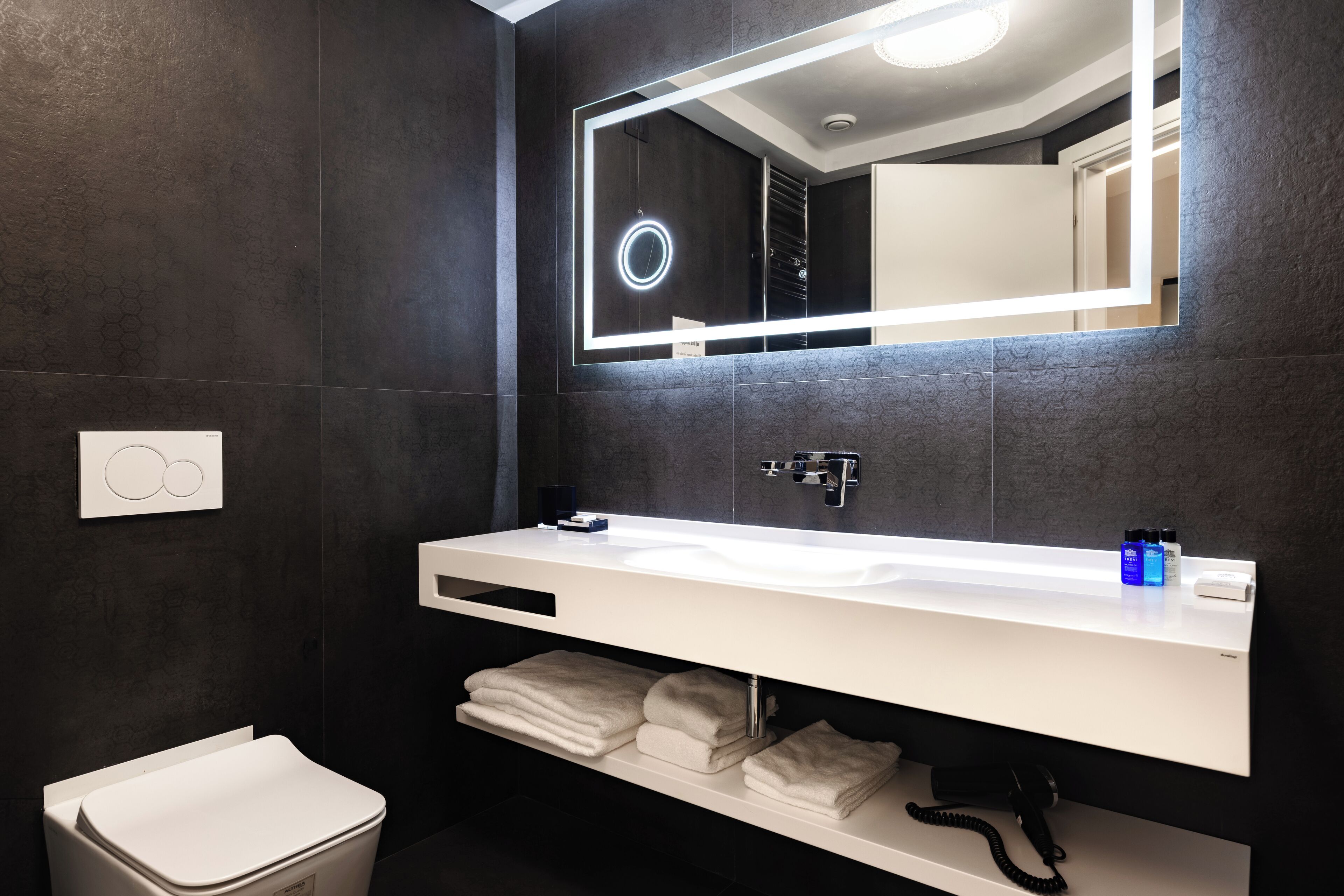 Een moderne badkamer met een witte wastafel, donker betegelde wanden, een grote spiegel en een toilet.