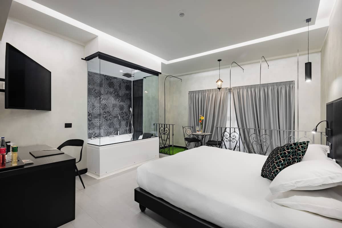Una habitación de hotel moderna con una cama grande, un escritorio con una silla, una mesita con una silla, un televisor montado en la pared y una ventana con cortinas.