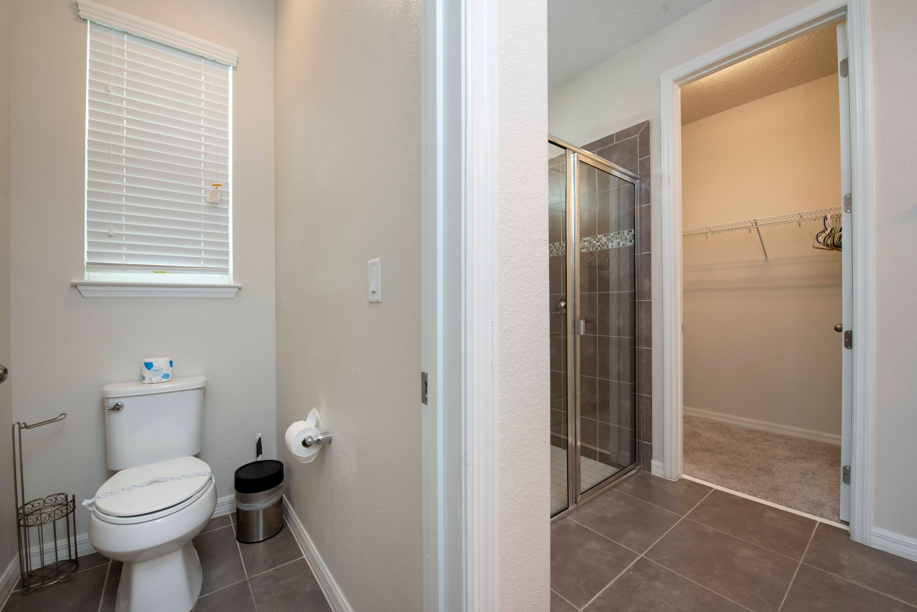 A bathroom with a toilet, a trash can, a window with blinds, and a shower area with a glass door.