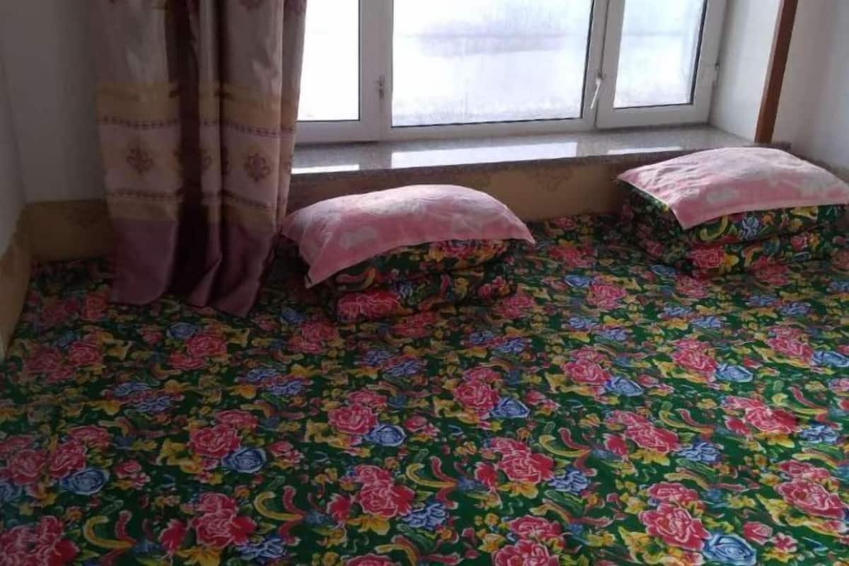 A room with a floral-patterned bedspread, a window with curtains, and a small bedside table.