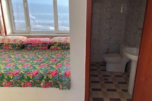 A bedroom with a colorful floral bedspread, a window with a view, and a bathroom with a toilet and sink. - Mohe Yujia Inn (Mohe)