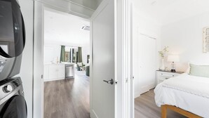 A hotel room with a bed, a nightstand, a lamp, and a view into the kitchen area. - Flourish at The Cove Riverwalk Villas (Wilmington)