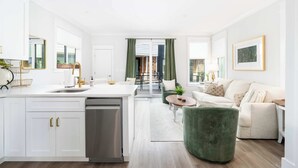 A modern kitchen with white cabinetry, a dishwasher, and a dining area with a green ottoman and a white sofa. - Flourish at The Cove Riverwalk Villas (Wilmington)