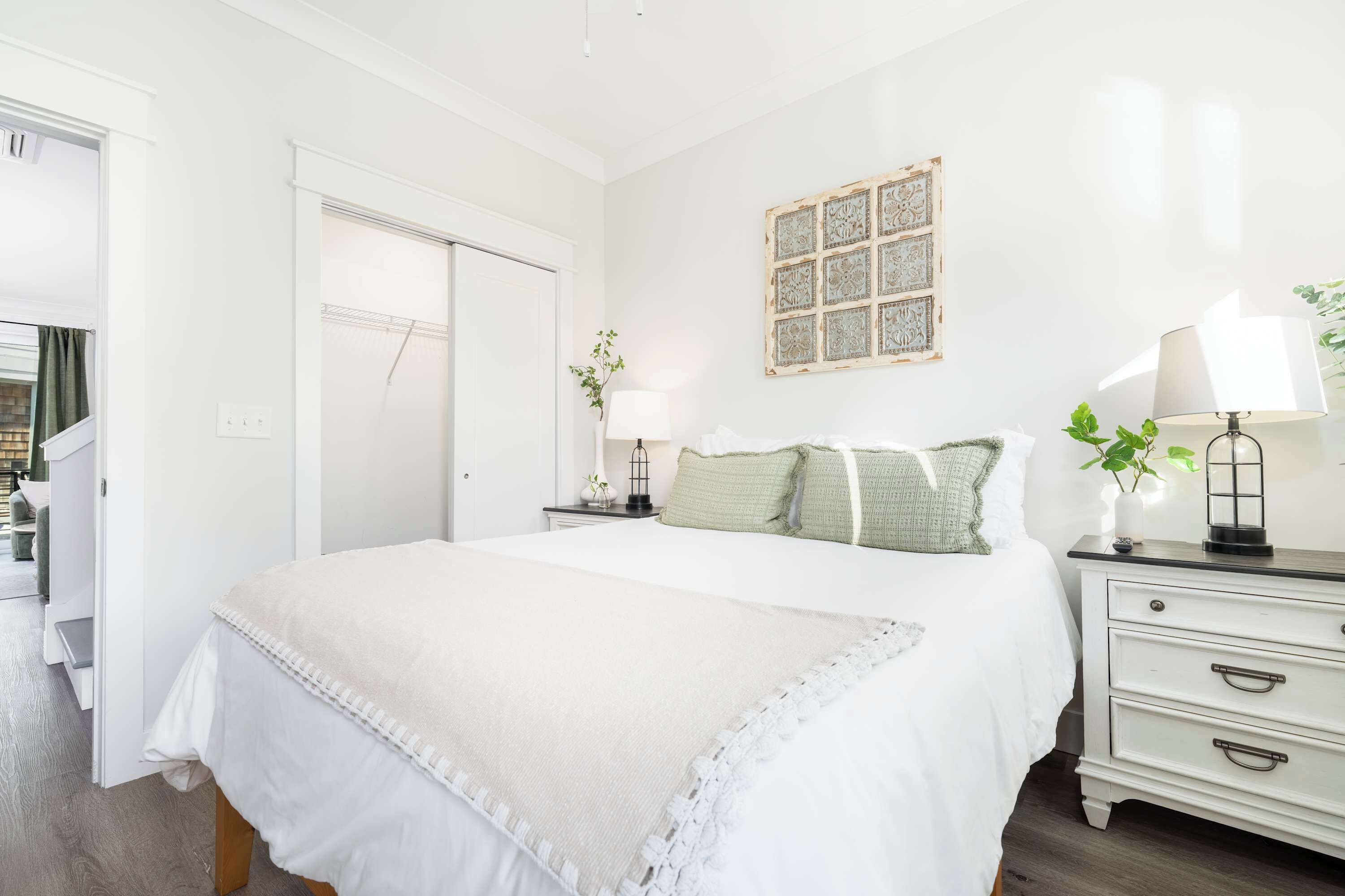Un dormitorio con cama, mesitas de noche, cómoda, puerta y una obra de arte colgada en la pared.
