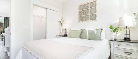 Un dormitorio con cama, mesitas de noche, cómoda, puerta y una obra de arte colgada en la pared.