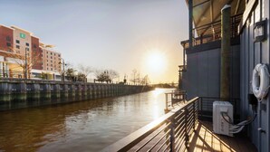 A waterfront hotel with a view of the river, a wooden deck, and a building with a visible 'E' sign. - Flourish at The Cove Riverwalk Villas (Wilmington)