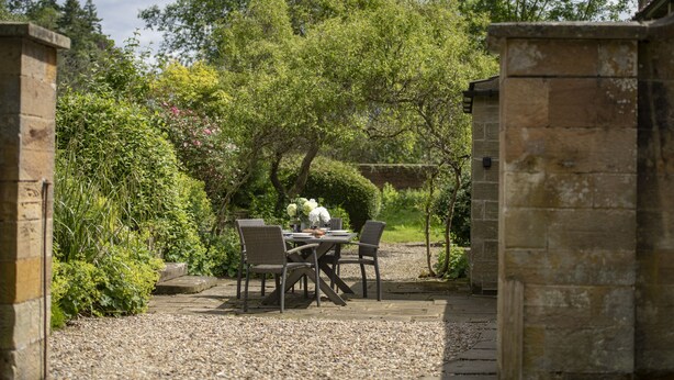 Une terrasse de jardin avec une table dressée pour quatre personnes, entourée de murs en pierre et d’une végétation luxuriante.