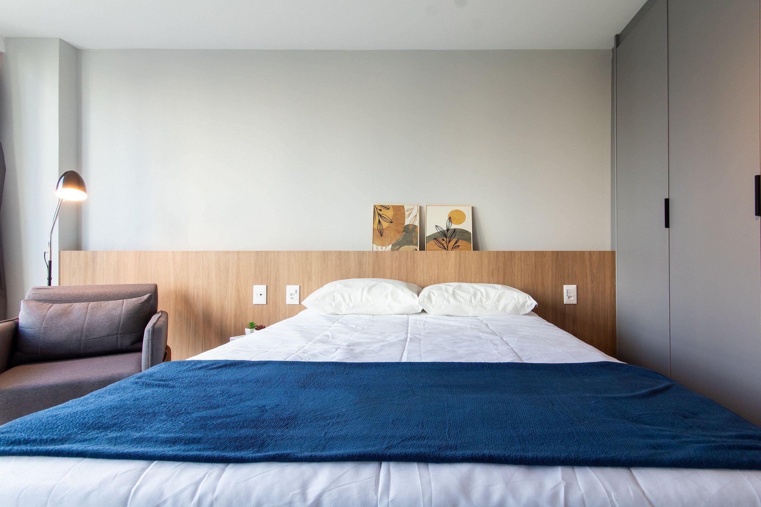 A hotel room with a bed, a chair, a lamp, and artwork on the wall.
