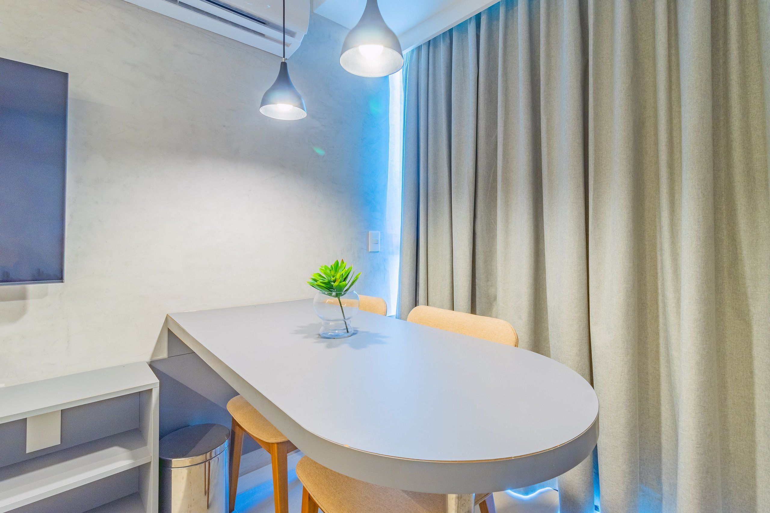 A modern meeting room with a white table, two pendant lights, a potted plant, and a large window with curtains.