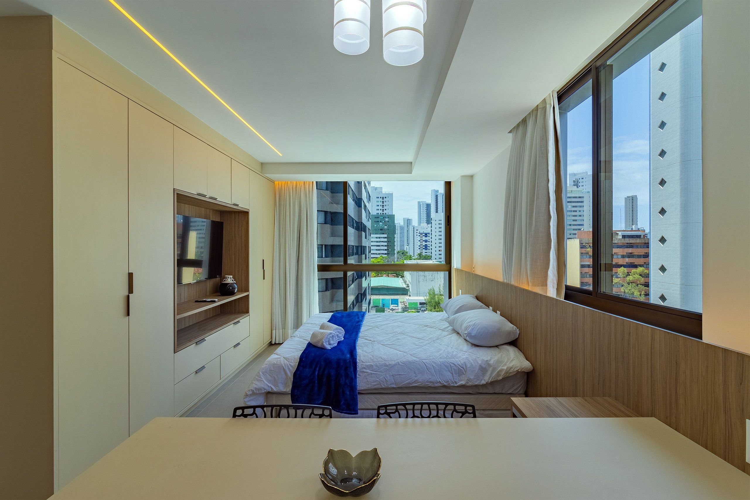 A modern hotel room with a bed, a TV, a dining table, and a view of the cityscape.