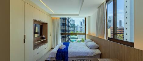 A modern hotel room with a bed, a TV, a dining table, and a view of the cityscape.