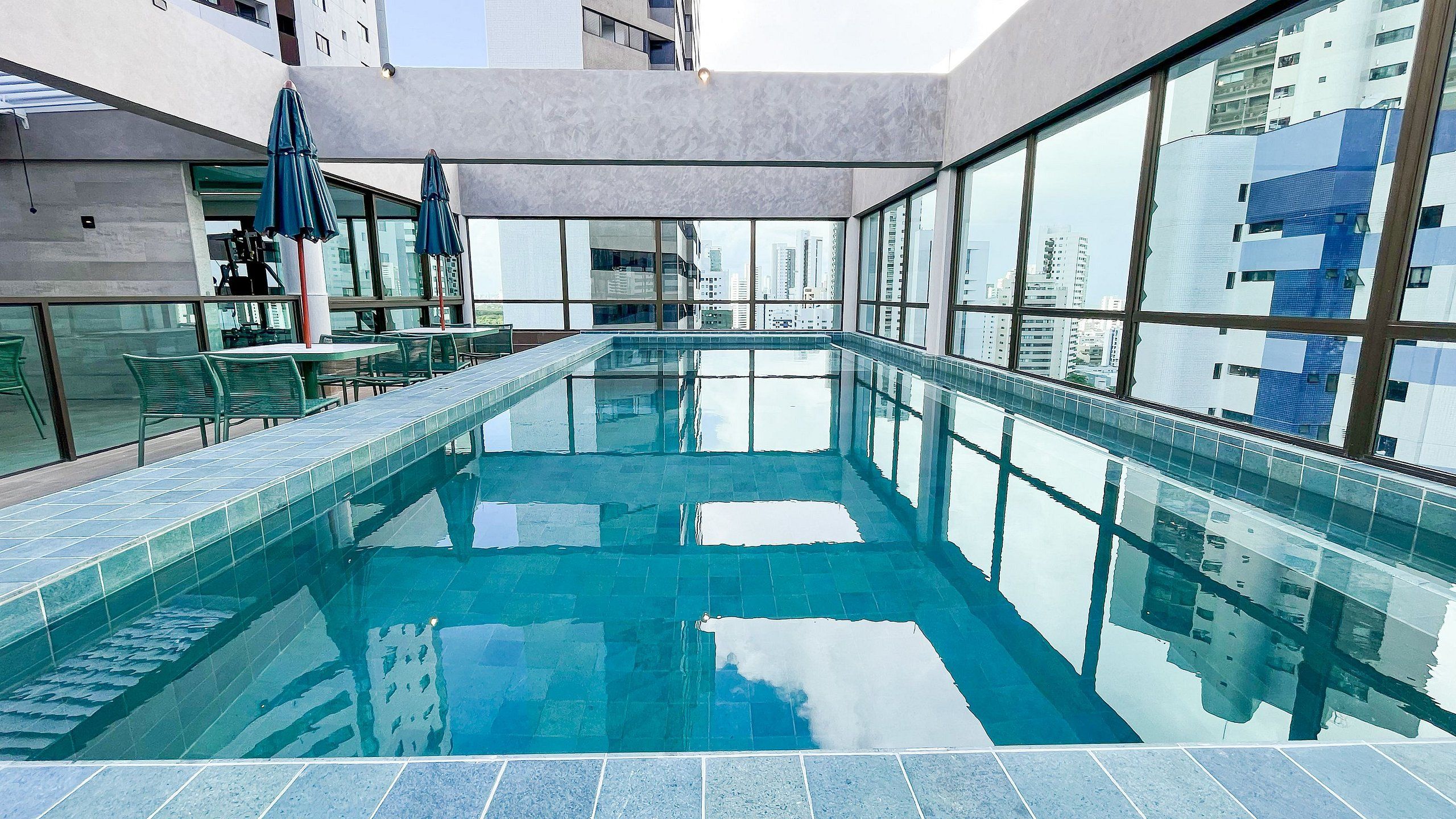 Piscina interior moderna com paredes de vidro transparente, piso ladrilhado e vista para edifícios da cidade.