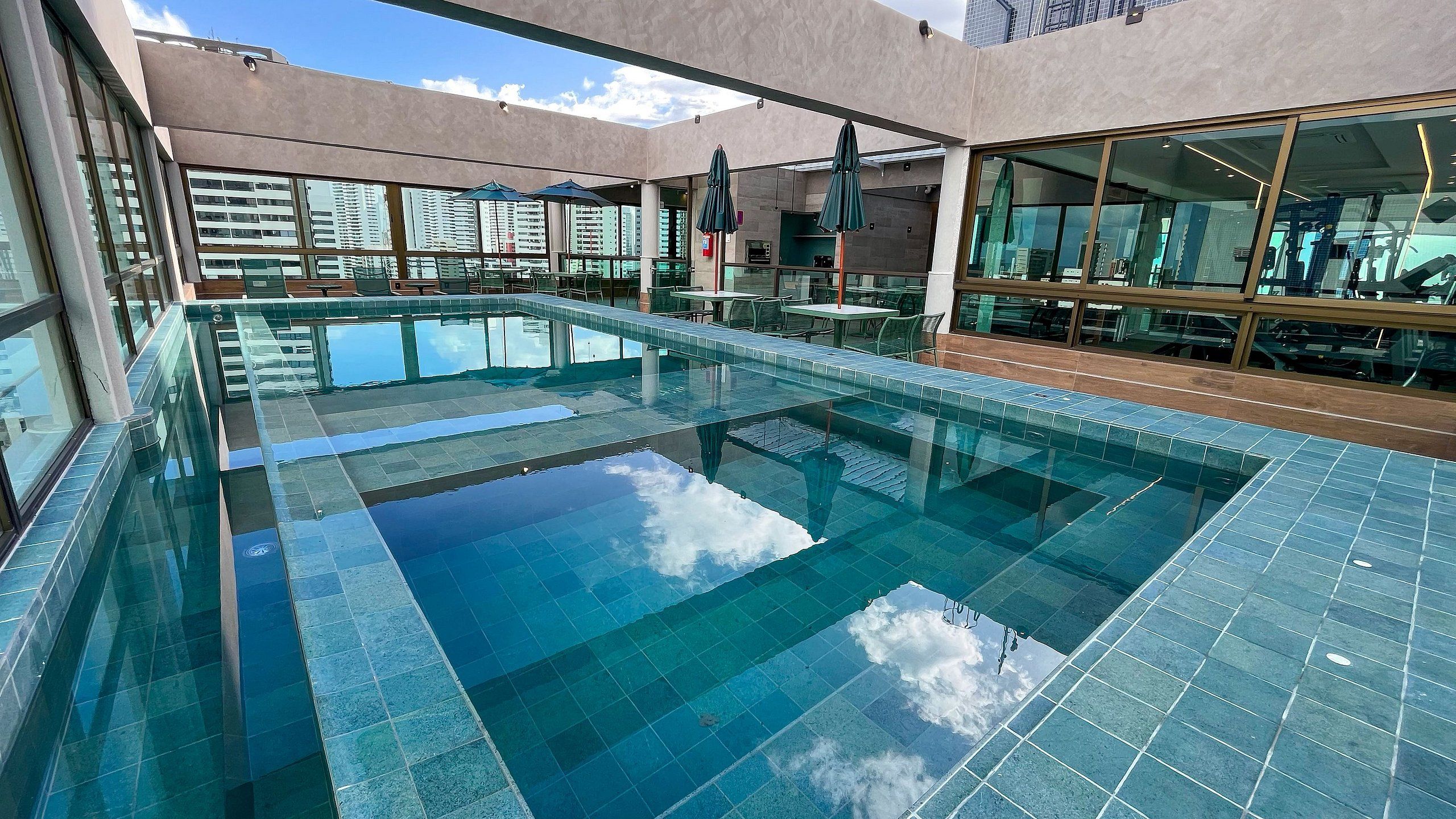 A modern indoor swimming pool with clear blue water, surrounded by glass walls and windows, offering views of the cityscape.