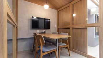 Uma área de refeições com mesa e cadeiras de madeira, uma TV de tela plana na parede e uma janela grande.