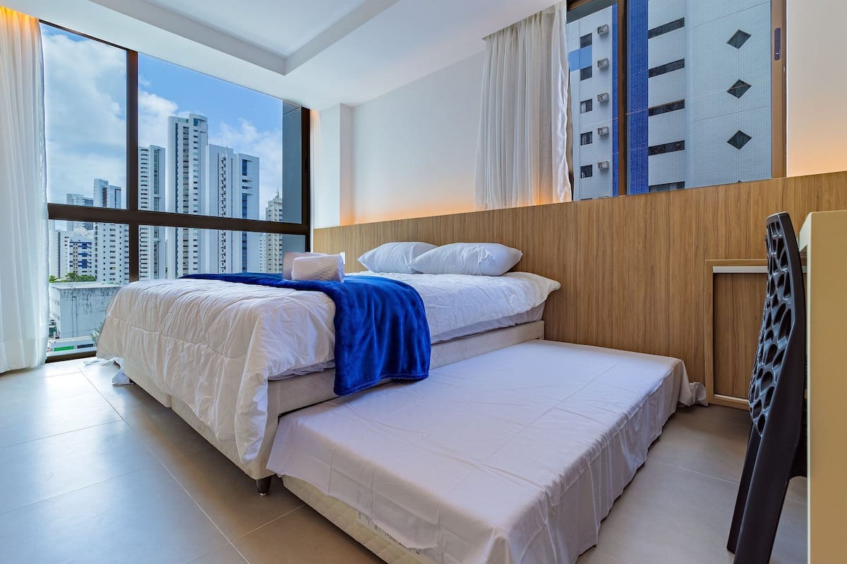 A hotel room with a large window, a bed, a wooden headboard, and a view of tall buildings.