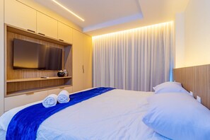 Une chambre d’hôtel moderne dotée d’un grand lit, d’un téléviseur à écran plat fixé sur un panneau en bois, d’une table de chevet avec une lampe et d’une grande fenêtre avec des rideaux.