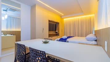 Quarto de hotel moderno com cama, mesa de jantar com cadeiras, televisão e cozinha compacta.