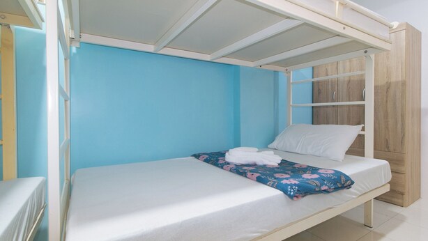 A bunk bed with a white mattress and a floral blanket, a white pillow, and a wooden wardrobe against a blue wall. - Catlea Inn (Pasay)