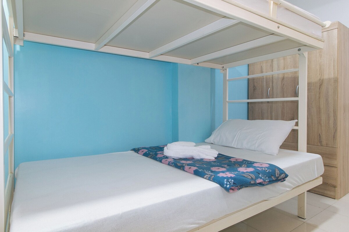 A bunk bed with a white mattress and a floral blanket, a white pillow, and a wooden wardrobe against a blue wall.