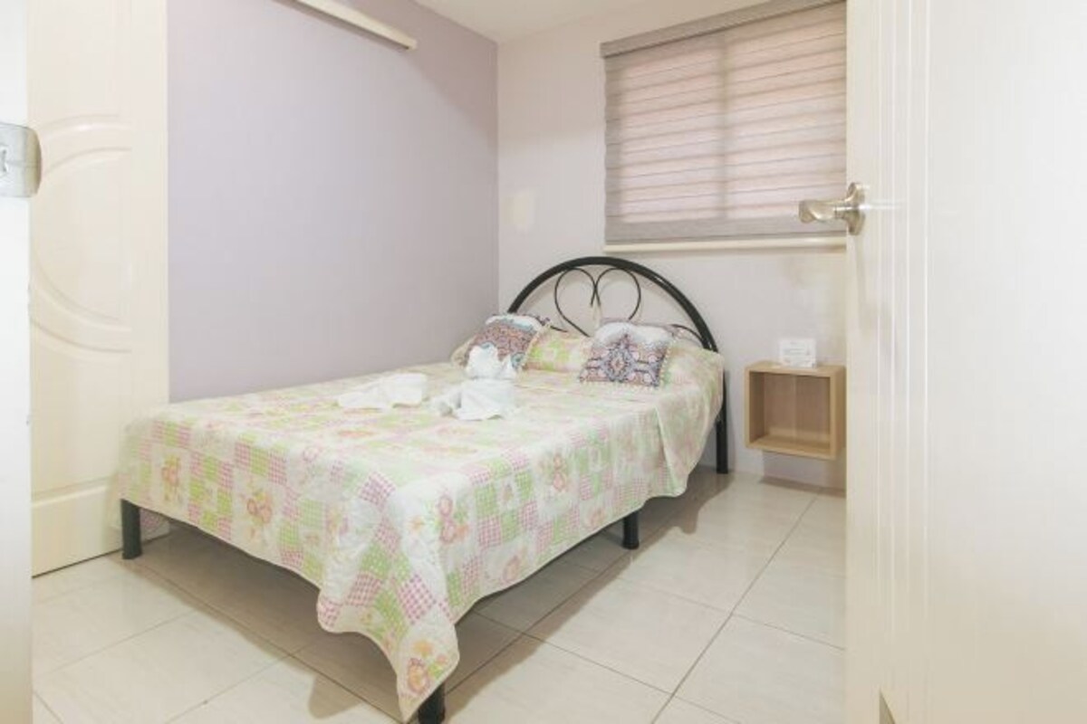 A small, neatly made bed with a floral bedspread in a room with light-colored walls and a window with blinds.