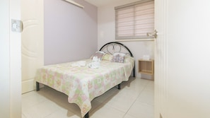 A small, neatly made bed with a floral bedspread, a bedside table with a lamp, and a window with blinds. - Catlea Inn (Pasay)