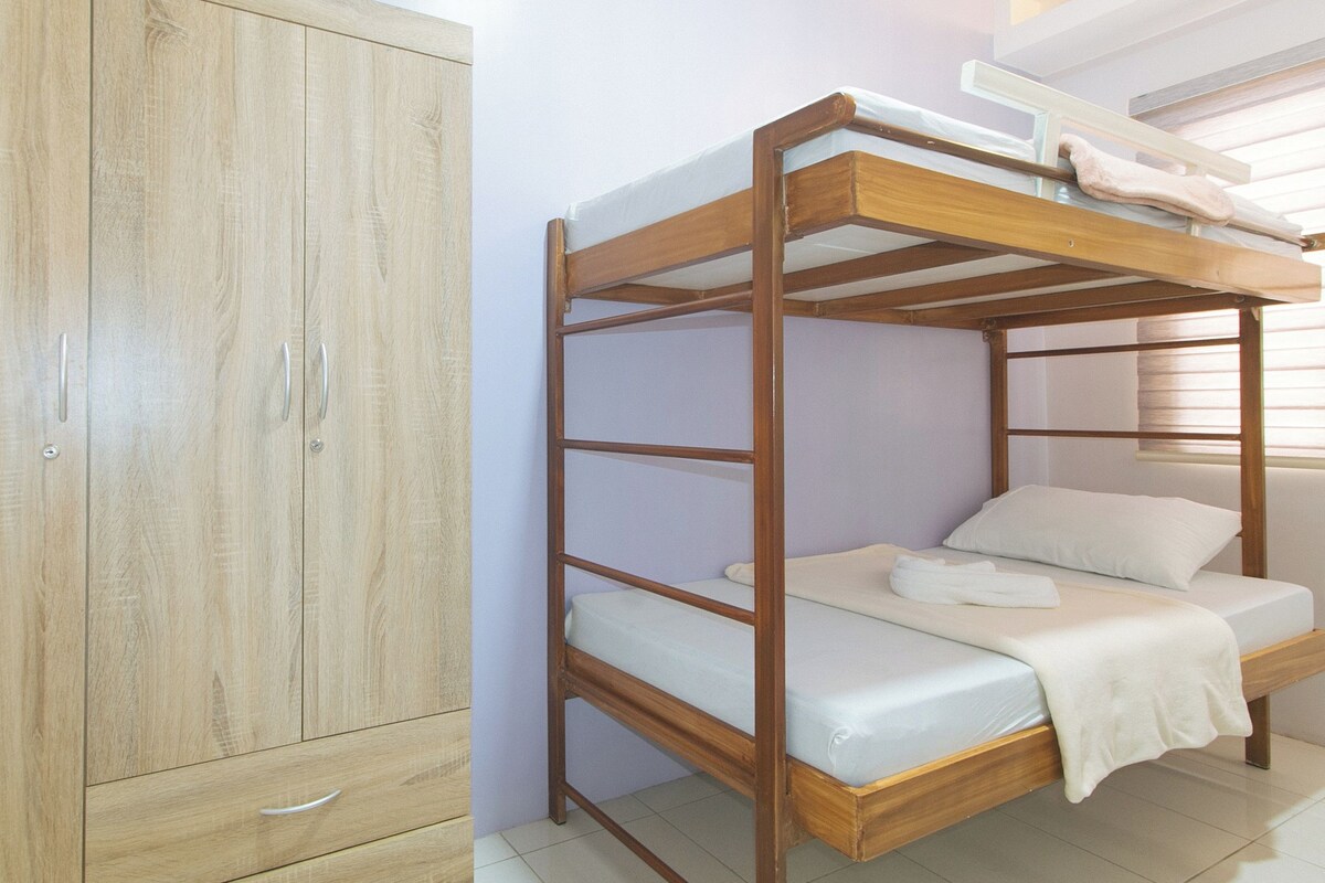 A bunk bed with a wooden frame and white bedding, a wooden wardrobe with two doors and a drawer, and a window with blinds.