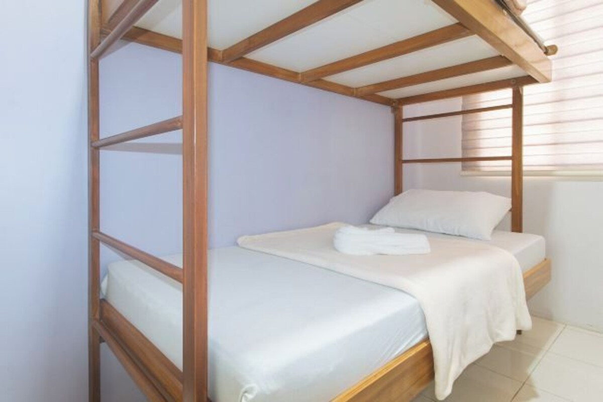 A single bed with a wooden frame and white bedding in a room with light-colored walls and a window with blinds.