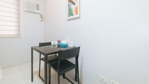 A small, clean room with a table and chairs, a window with blinds, a wall-mounted air vent, and a framed picture.