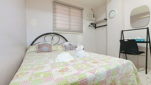 A neatly made bed with a floral quilt, a bedside table with a lamp, a chair, and a wall-mounted shelf with a mirror and clock.
