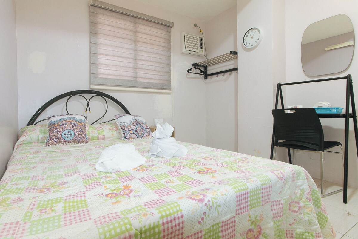 Un lit bien fait, recouvert d’une courtepointe à motifs floraux, une table de chevet avec une lampe, une chaise et une étagère murale agrémentée d’un miroir et d’une horloge.