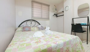 Un lit bien fait, recouvert d’une courtepointe à motifs floraux, une table de chevet avec une lampe, une chaise et une étagère murale agrémentée d’un miroir et d’une horloge.