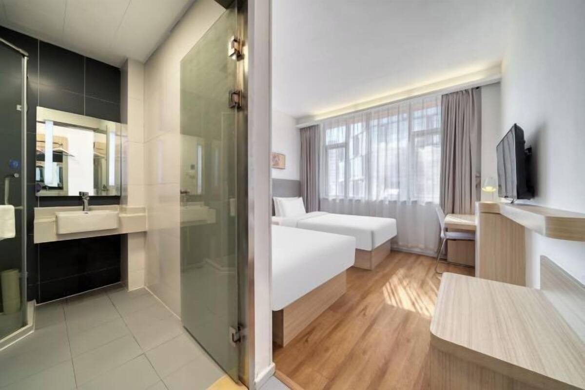 A modern hotel room with a glass shower enclosure, a large bed with a wooden headboard, a bedside table, and a TV mounted on the wall.