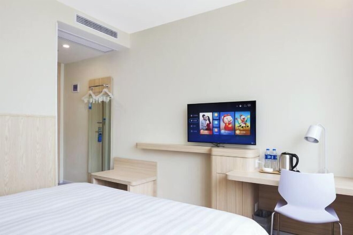 A hotel room with a bed, a TV on the wall, a desk with a lamp, and a shower area.