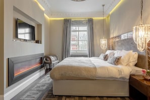 A hotel room with a bed, a TV mounted on the wall, a heater, a window with curtains, and a chandelier.