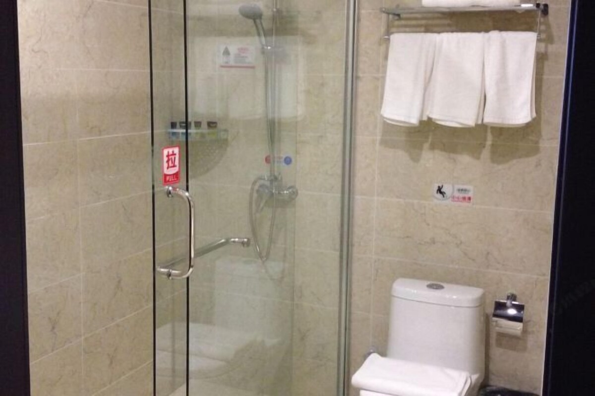 A glass-enclosed shower stall with a handrail, a toilet with a paper towel dispenser, and a towel rack with towels.