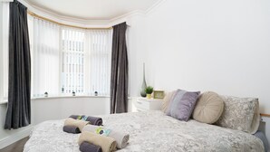 A neatly made bed with rolled towels, a bedside table with a plant, and a window with curtains. - Beautiful 4 Bedroom House Immaculate Condition. Just Refurbished House. (Romford)