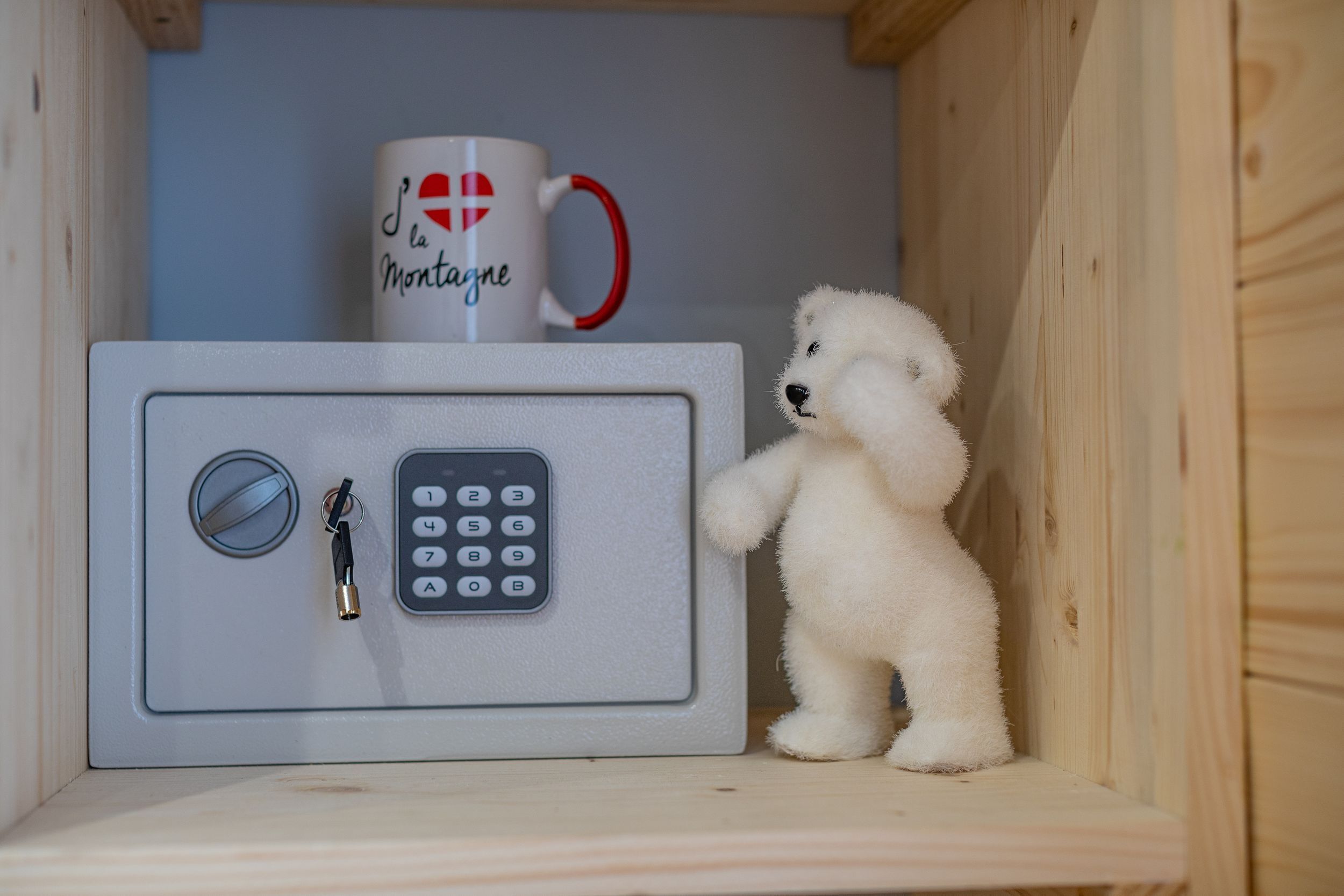 Ein kleiner, weißer Safe mit Zahlenschloss und Schlüsselloch, daneben ein Teddybär und eine Tasse mit Herz und Schriftzug.