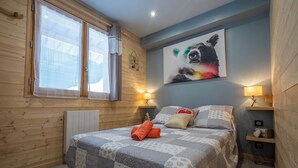 Ein Schlafzimmer mit Holzboden, einem Bett mit grauer und weißer Steppdecke, zwei Nachttischlampen, einem Wandbild eines Bären und einem Fenster mit Vorhängen.