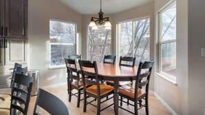 A dining room with a wooden table and chairs, large windows, and a chandelier.
