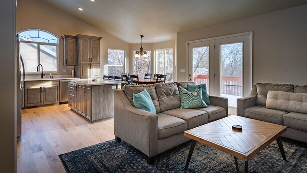 A spacious living area with a large sofa, a coffee table, and a dining table with chairs. There is a kitchen with wooden cabinets and a large window.