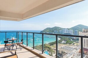 A balcony with a view of the sea and mountains, a table with chairs, and a glass railing.