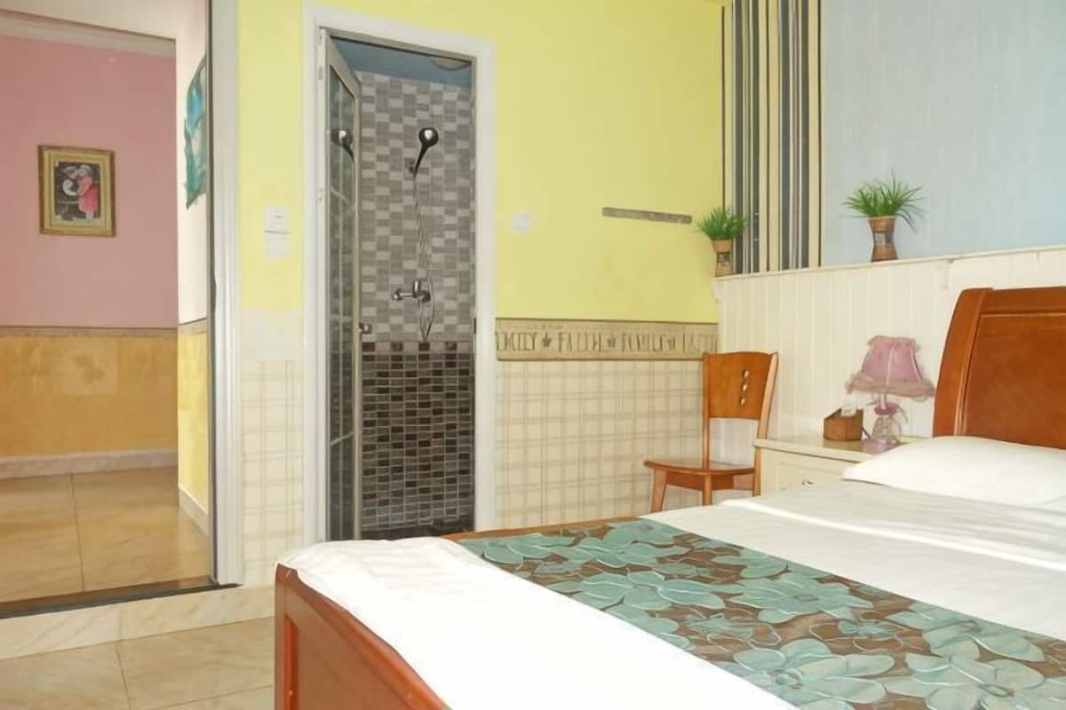 Een slaapkamer met een bed, een stoel, een nachtkastje, een lamp, een potplant en een douche.
